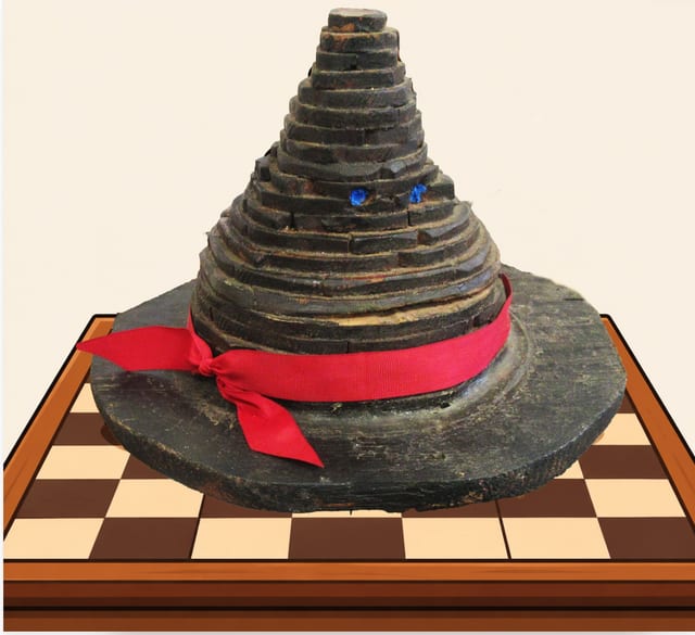 The talking hat: a tall witch&rsquo;s hat built up from rings of dark, rough-cut wood, with two small blue eyes peering out and a red ribbon tied in a bow round its brim, standing on a drawn chessboard.